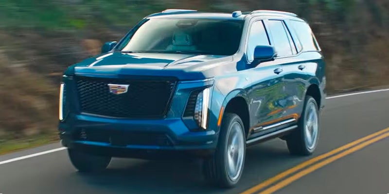 Blue Cadillac SUV driving on a two-lane road, featuring a prominent front grille and vertical LED headlights