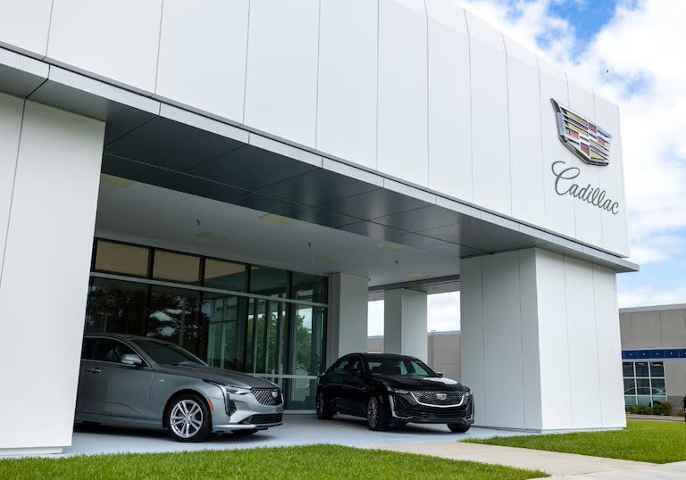 Exterior view of a Cadillac dealership with two Cadillac vehicles parked under a covered entrance beside a glass-front showroom